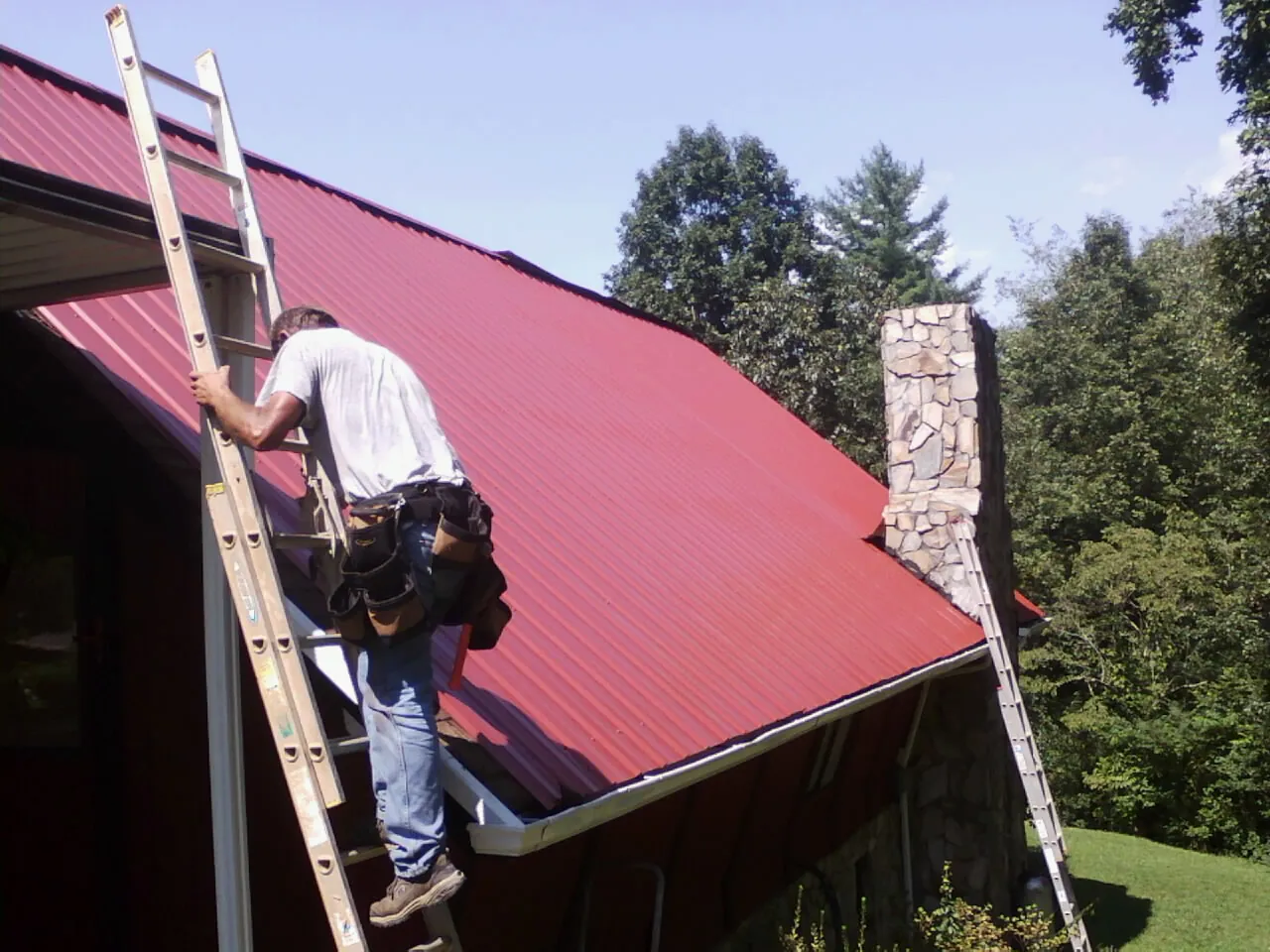 Roofer on a ladder beside a red metal roof and stone chimney — Soffit & Fascia Repair by Roofing Brothers in Vaughn